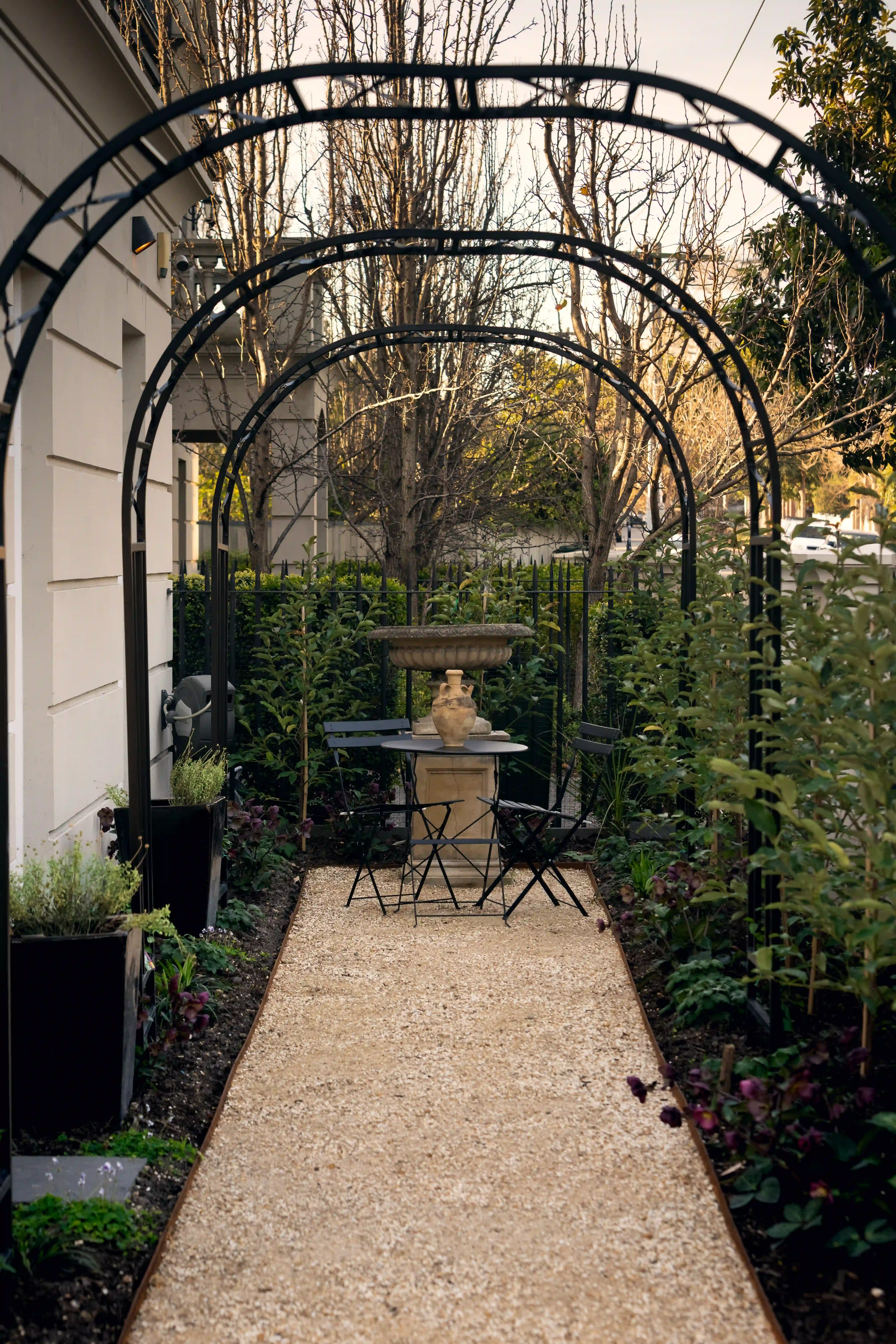 Gravel path running beneath a row of black metal garden arches toward a stone urn and black bistro setting.