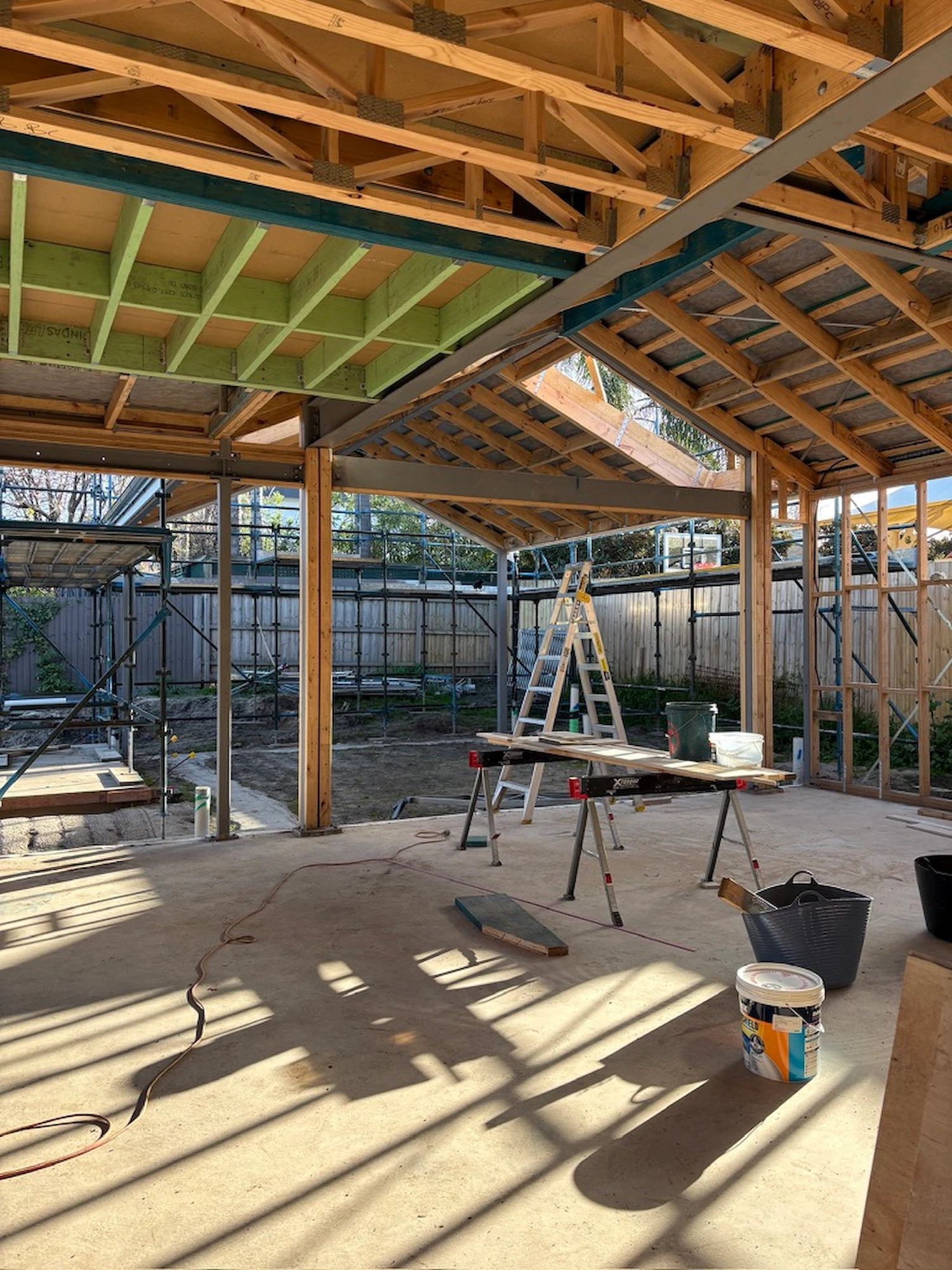 Timber-framed alfresco under construction at a Black Rock home, with exposed roof trusses and steel beams, a ladder and trestle, looking out to the garden beyond.