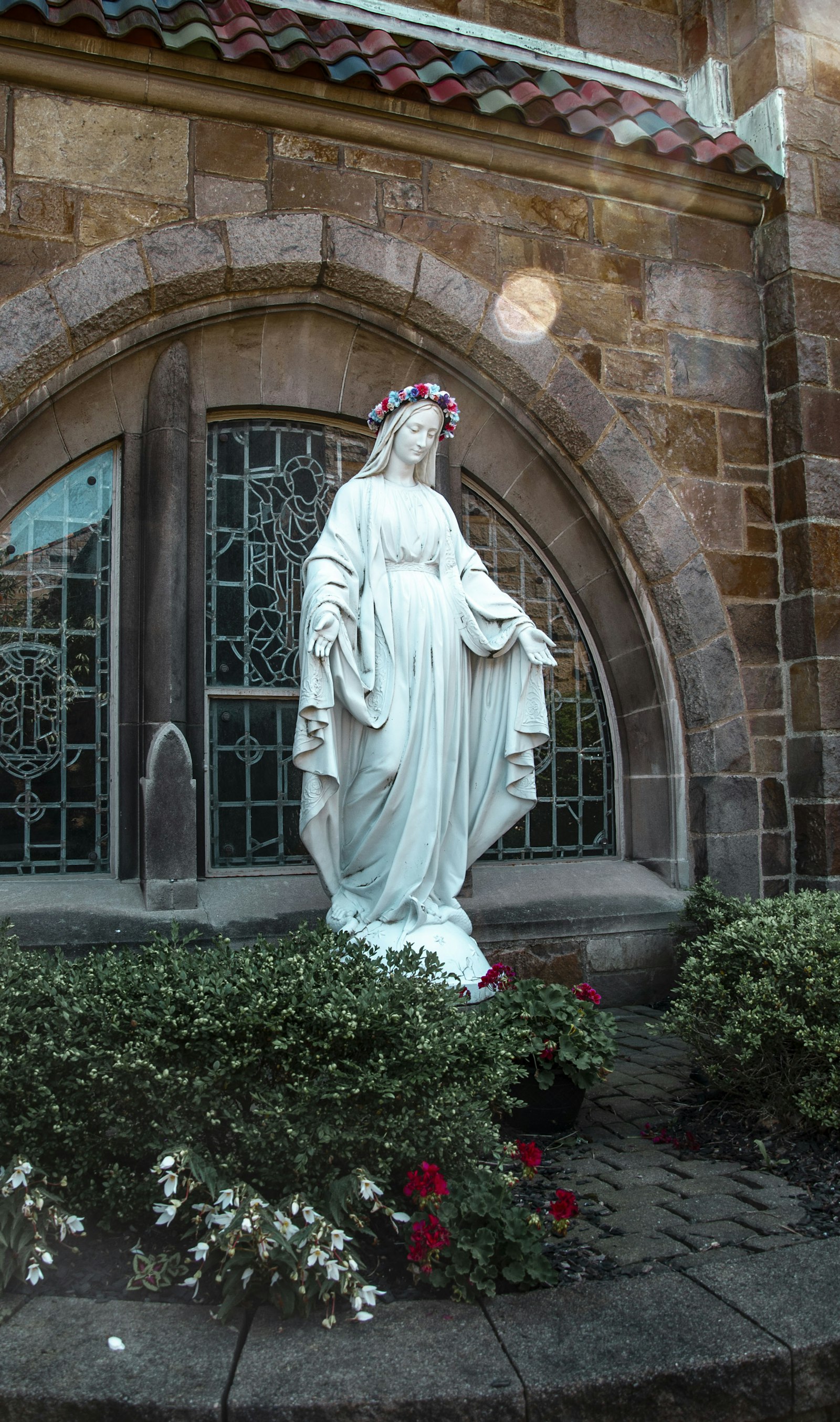 Patung Bunda Maria di halaman gereja batu, dikelilingi bunga dan tanaman hijau