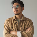 Man with glasses wearing a brown jacket and beige shirt, standing with arms crossed against a plain background.