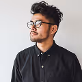 Man with dark hair and glasses wearing a black button-up shirt looking left against a white background.