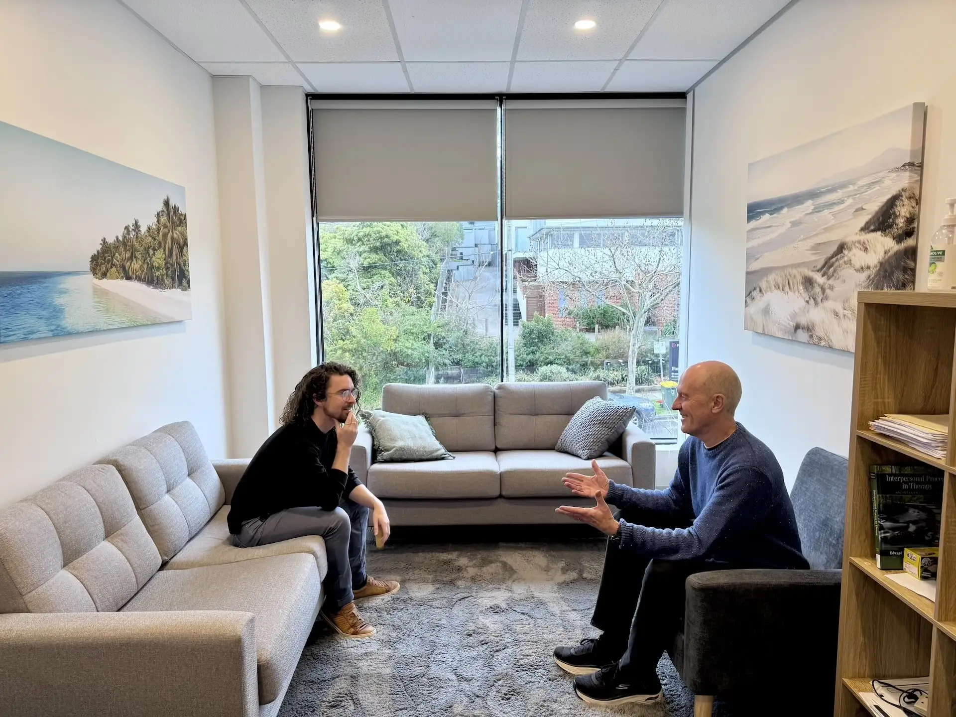Two men seated and engaged in conversation in a modern office lounge with gray sofas, a large window with blinds showing greenery outside, beach-themed wall art, and a bookshelf with books and hand sanitizer.