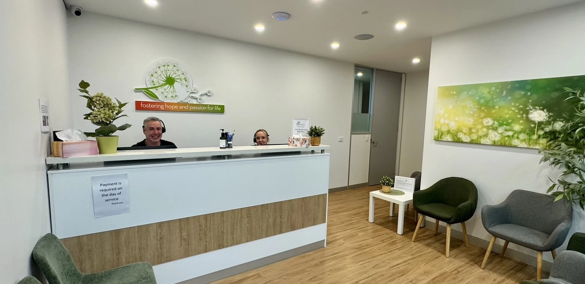 Modern reception area with two smiling staff wearing headsets behind a white and wood-paneled desk, decorated with plants and hand sanitizer; waiting area has gray and green chairs, a white table, a large dandelion-themed painting, wood flooring, and a sign on the desk reading 'Payment is required on the day of service.'