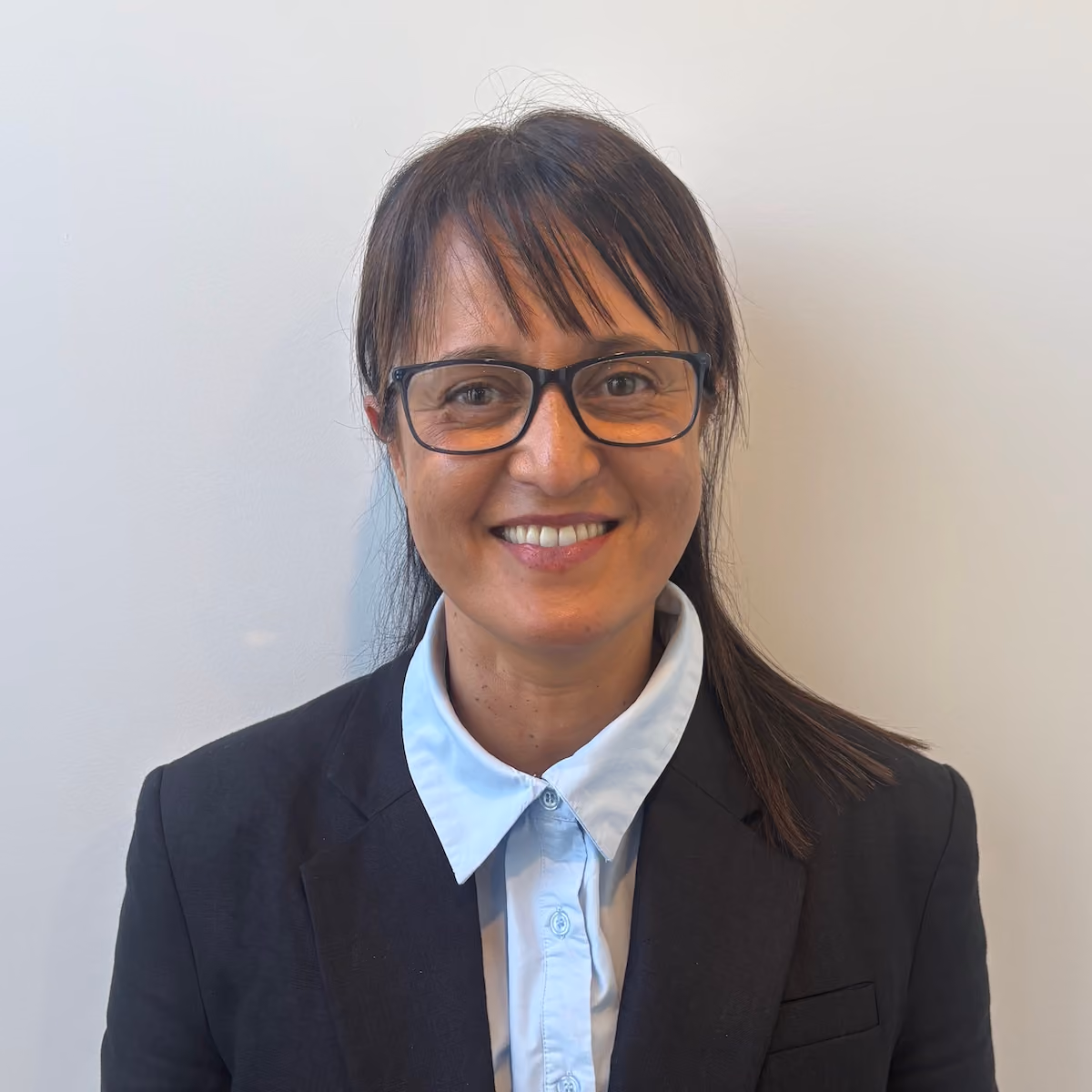 Smiling woman with straight dark brown hair and bangs, wearing glasses, a light blue collared shirt, and a black blazer, standing against a plain light background.