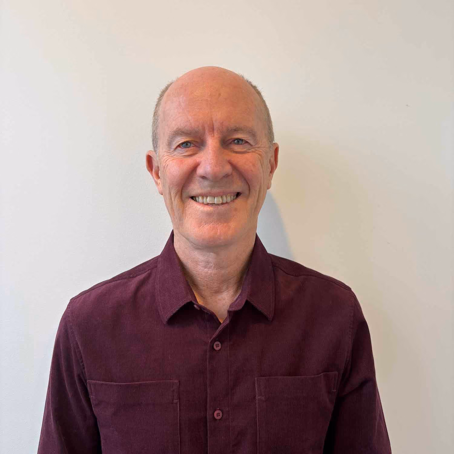 Smiling middle-aged man with a bald head wearing a dark maroon button-up shirt standing against a plain light-colored wall.