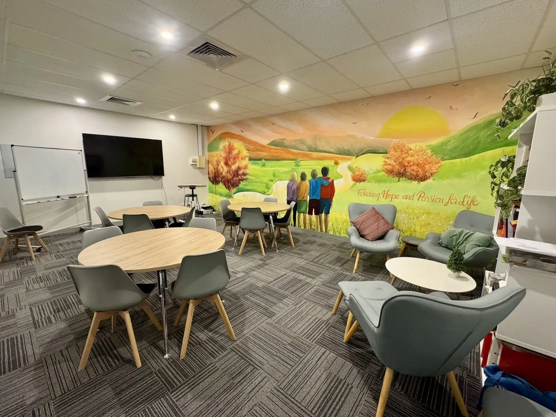 Bright room with round tables and gray chairs on carpeted floor, a wall mural showing four people standing arm in arm facing a green landscape with hills, trees, and a yellow sun, and the words 'Fostering Hope and Passion for Life.' There are also cushioned armchairs with pillows, a whiteboard, a large TV screen, and shelves with plants and books.