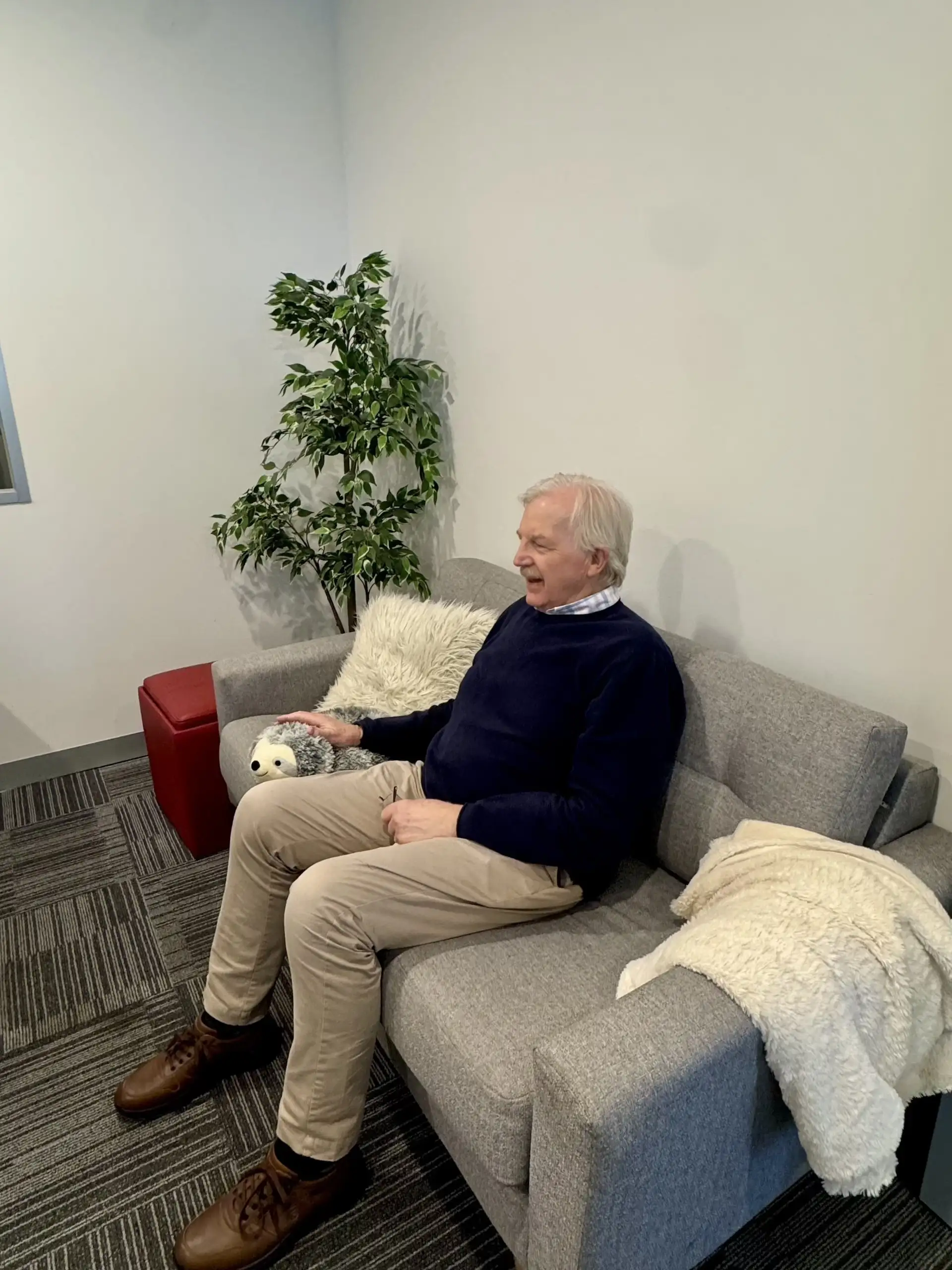 An elderly man wearing a navy blue sweater, beige pants, and brown shoes sits on a gray couch in a room with gray carpet and white walls, gently touching a small plush toy resembling a hedgehog. A white furry blanket is draped over the armrest, and a green leafy plant stands in the corner behind him.