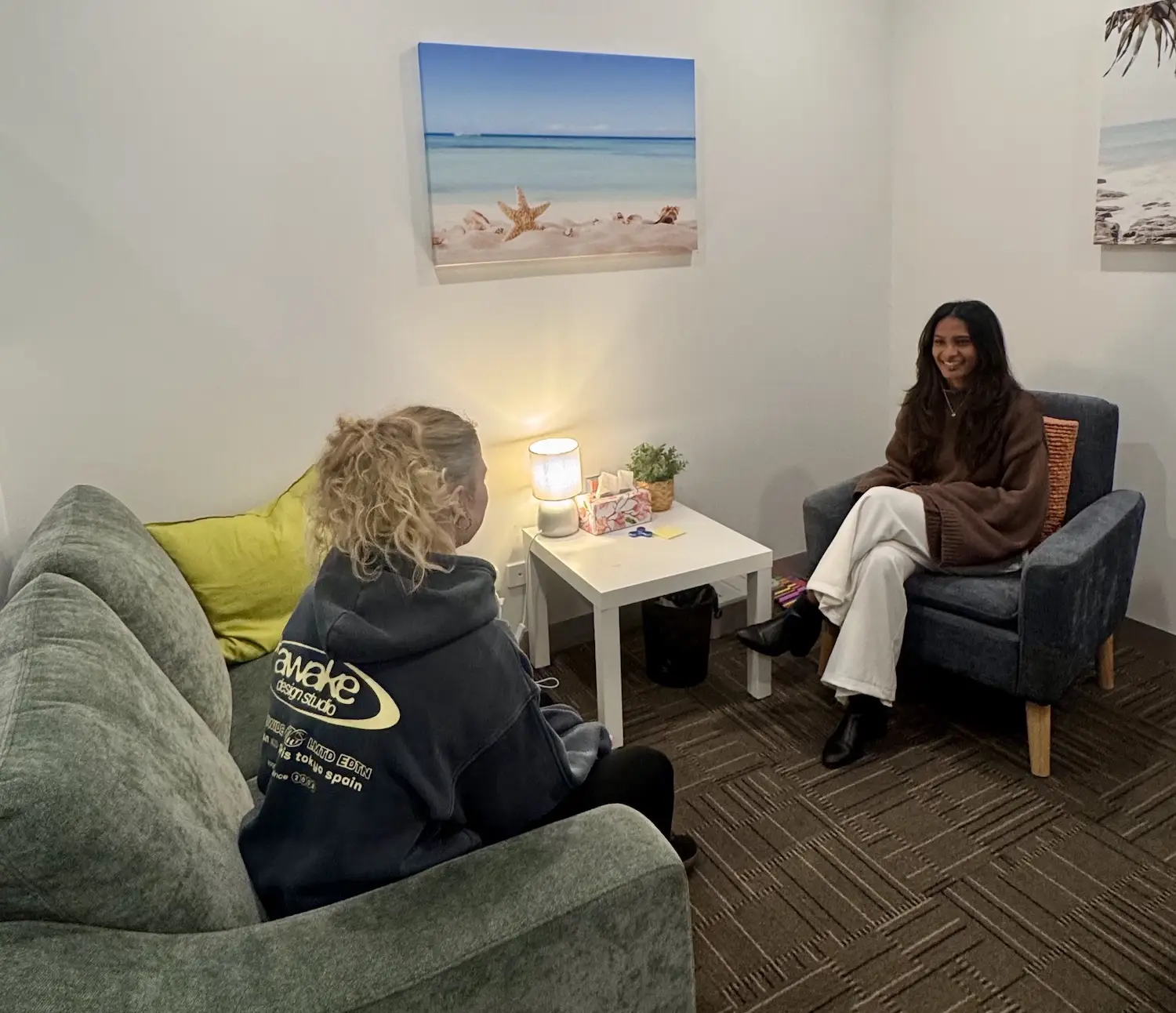Two women engaged in conversation in a cozy room, one sitting on a green sofa and the other on a gray armchair, with a small white table between them holding a lit lamp, tissue box, and a small plant, and beach-themed pictures on the walls.