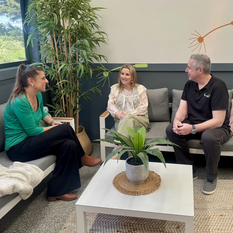 Three people, two women and one man, are seated and having a friendly conversation in a modern indoor lounge area with gray couches, a white coffee table with a potted plant, and a tall leafy plant in the corner near a large window.