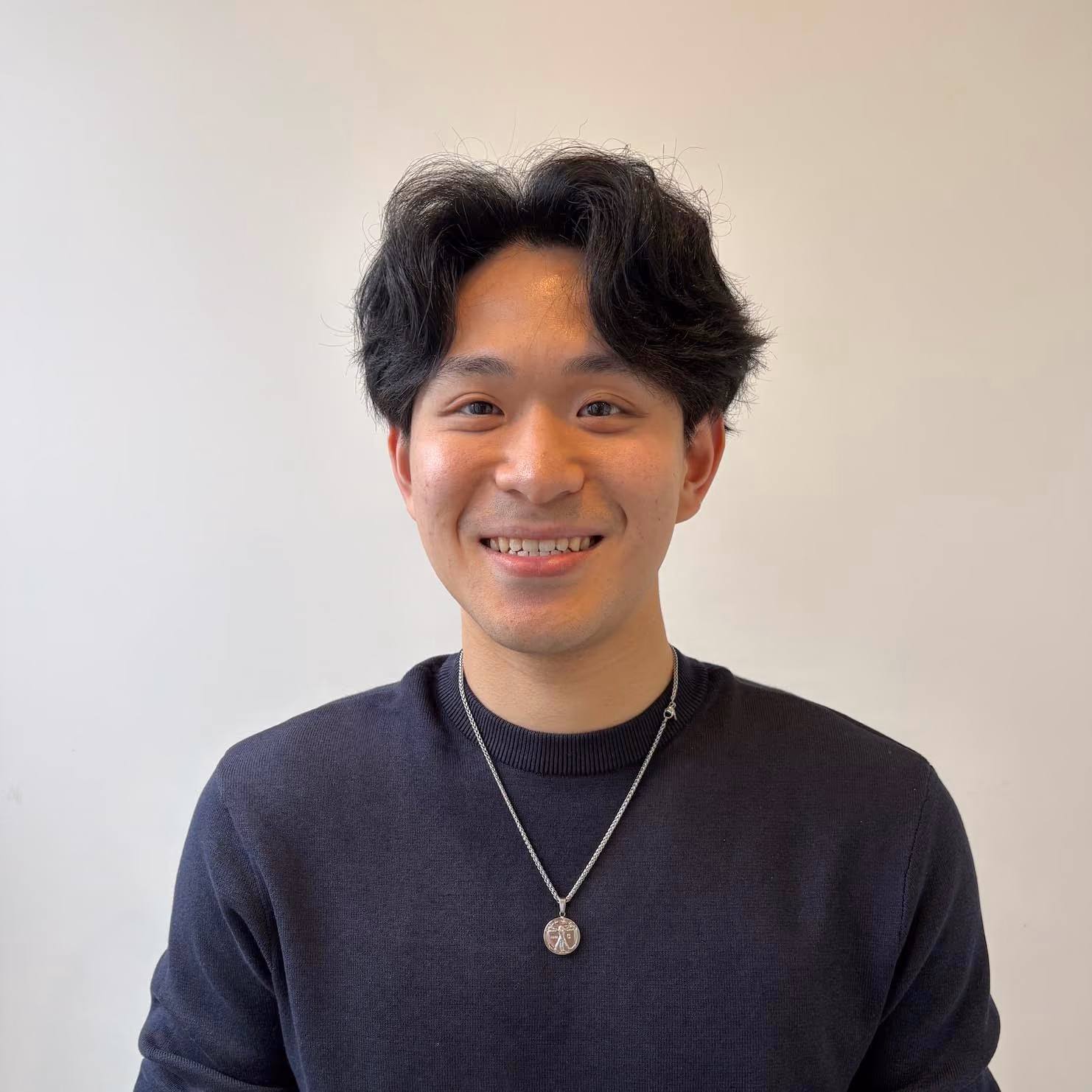 Smiling young man with wavy black hair wearing a dark navy sweater and a silver necklace with a pendant, posed against a plain light background.