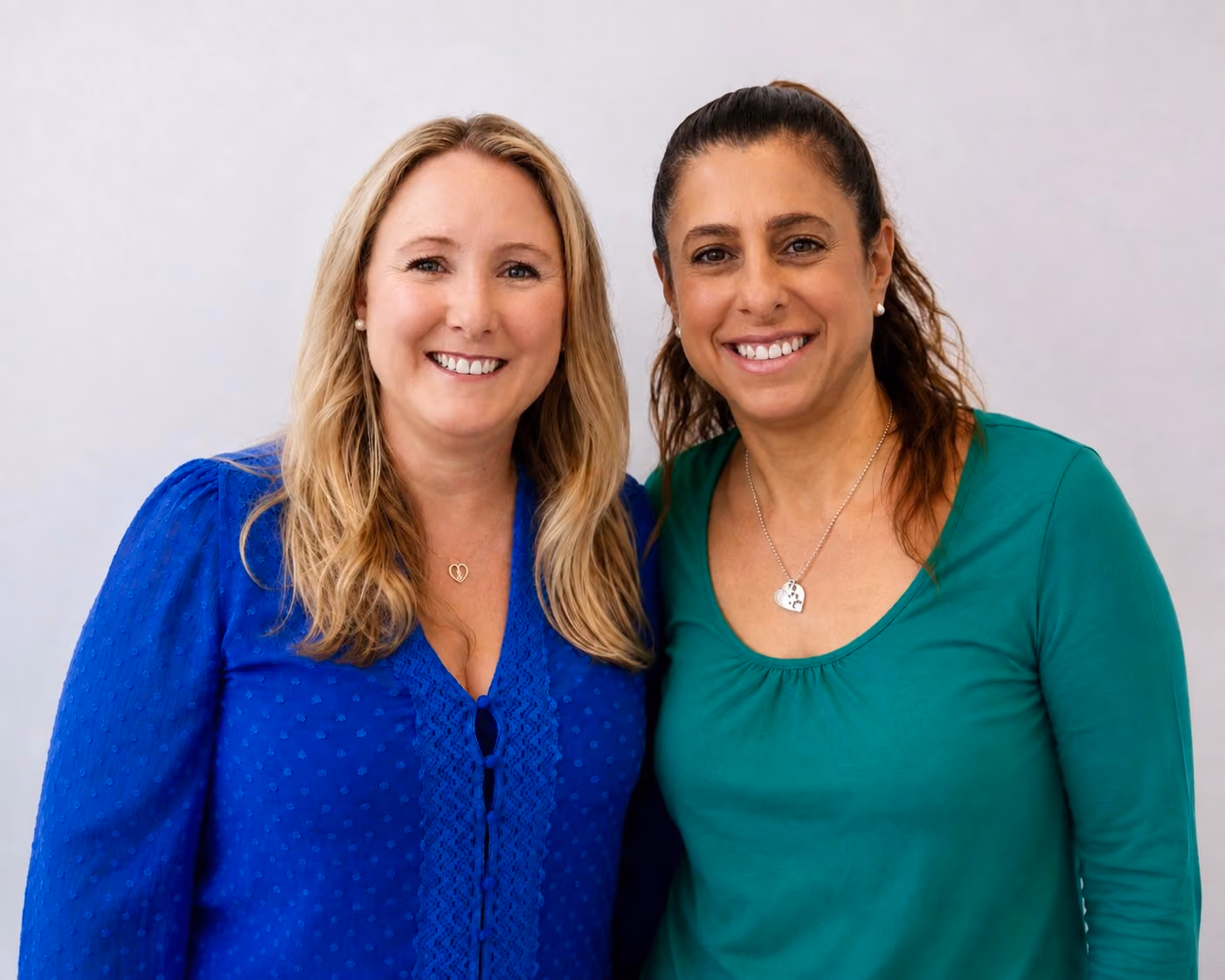Two women smiling and standing close together against a plain light background; the woman on the left has long blonde hair and wears a blue blouse, and the woman on the right has dark hair tied back and wears a green top with a necklace.