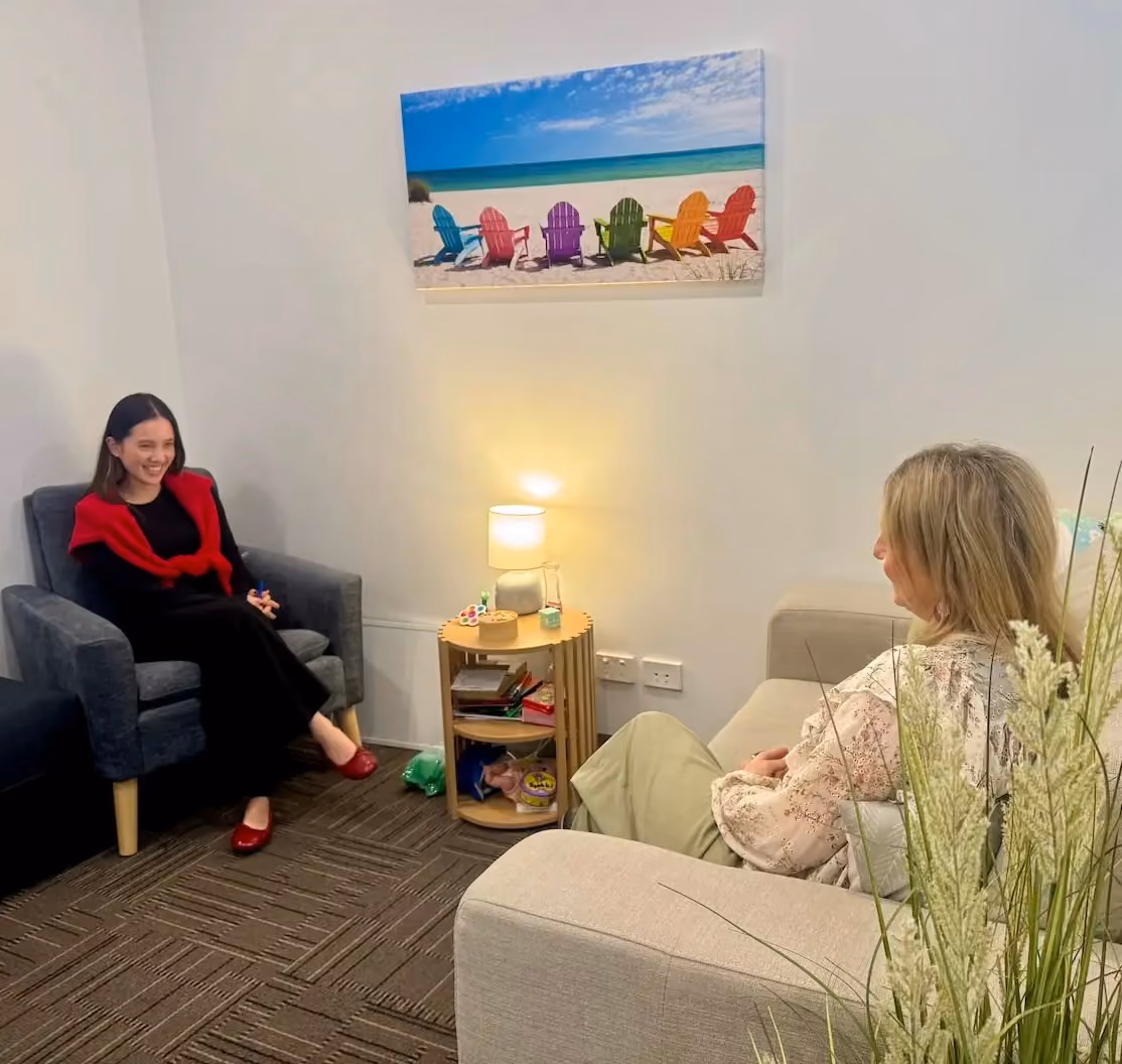 Two women sitting and conversing in a cozy room with an armchair, a sofa, a small round wooden side table with a lamp, and a colorful beach scene artwork on the wall.
