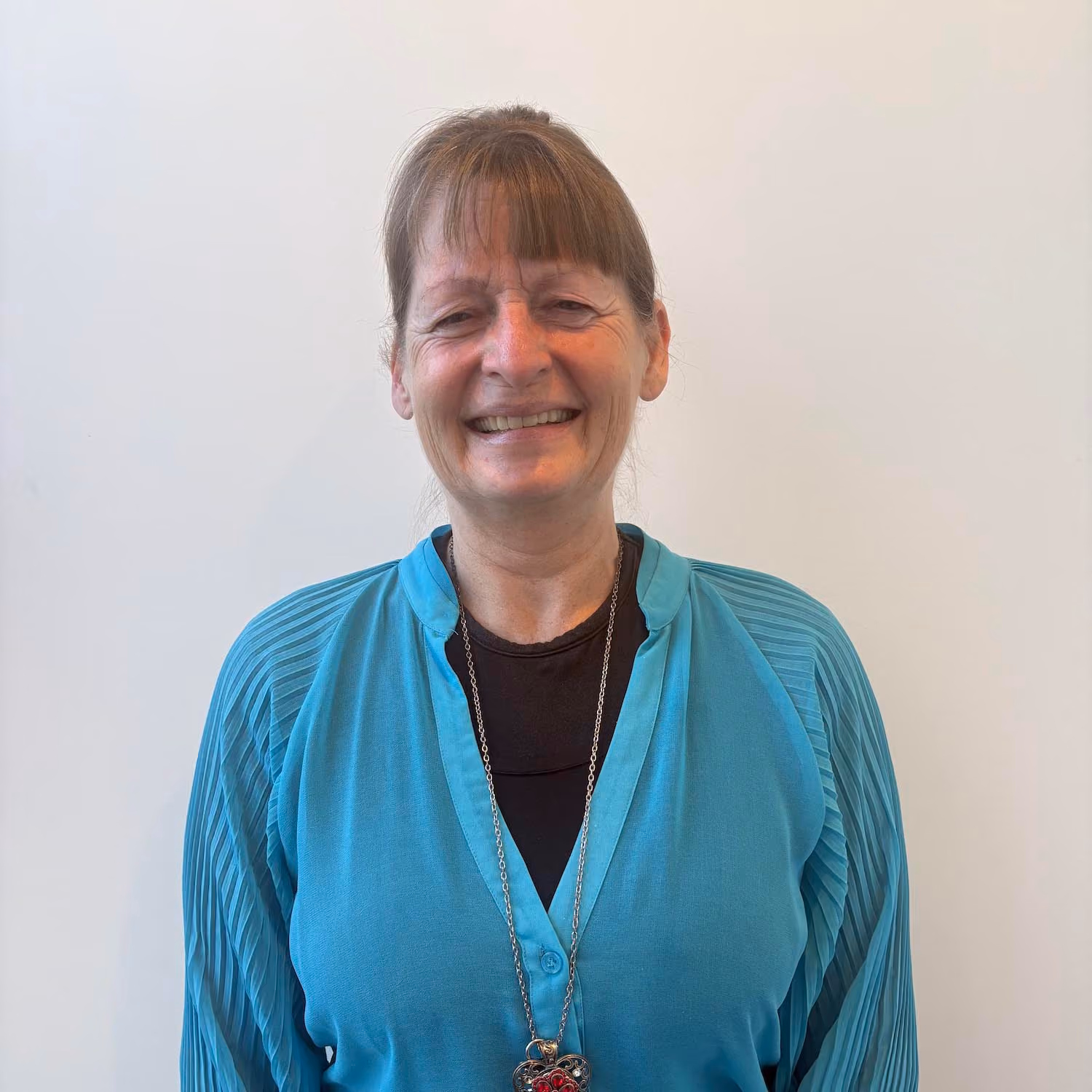 Smiling middle-aged woman with light brown hair tied back and bangs, wearing a turquoise cardigan over a black top, standing against a plain light background.