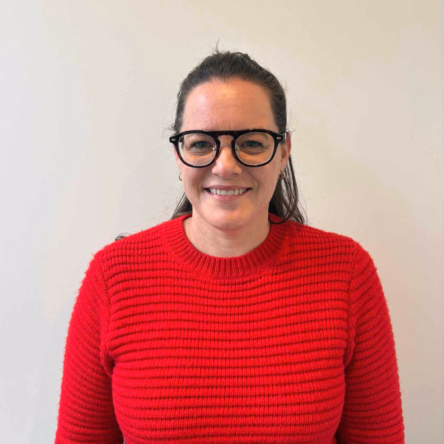 Smiling woman with dark hair tied back, wearing black-rimmed glasses and a red ribbed sweater, against a plain light background.