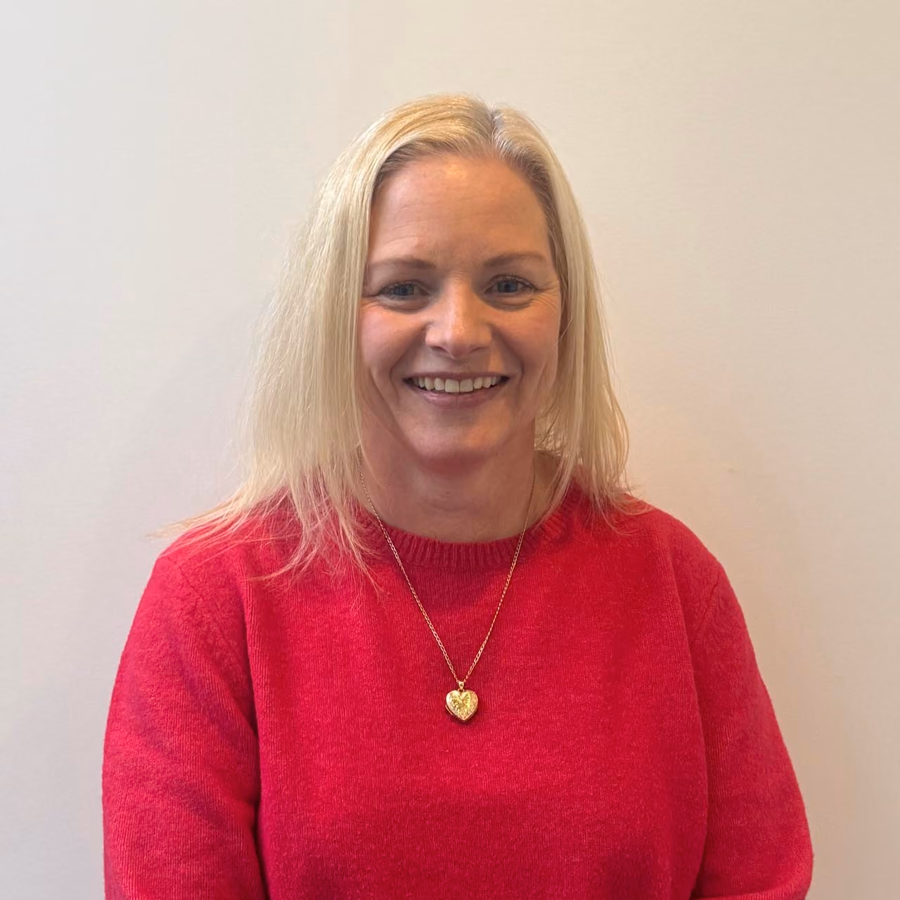 Smiling woman with straight blonde hair wearing a bright red sweater and a gold heart-shaped pendant necklace against a plain light background.