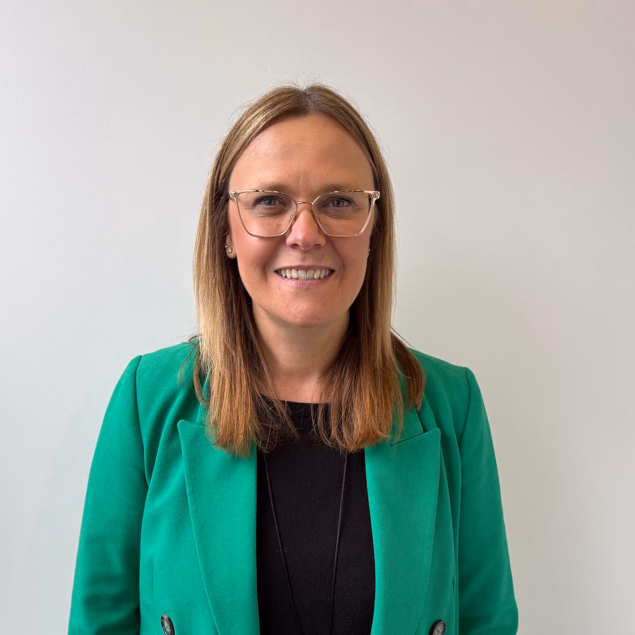 Smiling woman with straight, shoulder-length blonde hair wearing glasses, a green blazer, and a black top, standing against a plain light-colored background.