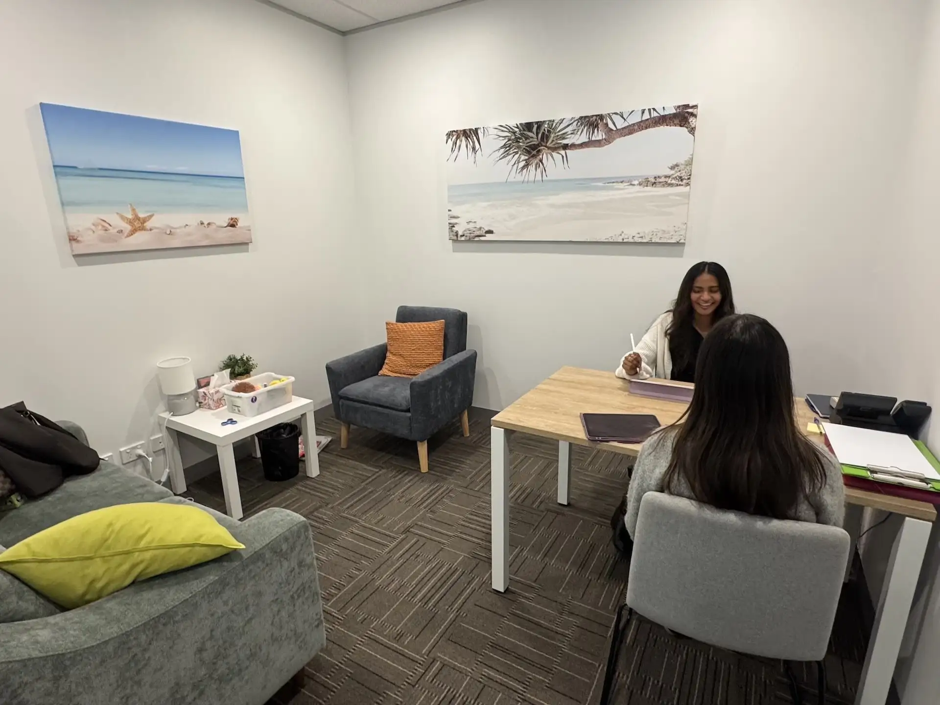 Two women in a small office; one woman is sitting behind a wooden desk smiling and holding a stylus with a tablet, the other is sitting on a gray chair facing her. The office has gray carpet, white walls with two beach-themed paintings, a gray armchair with an orange pillow, a gray couch with a yellow pillow, and a small white side table with items on it.