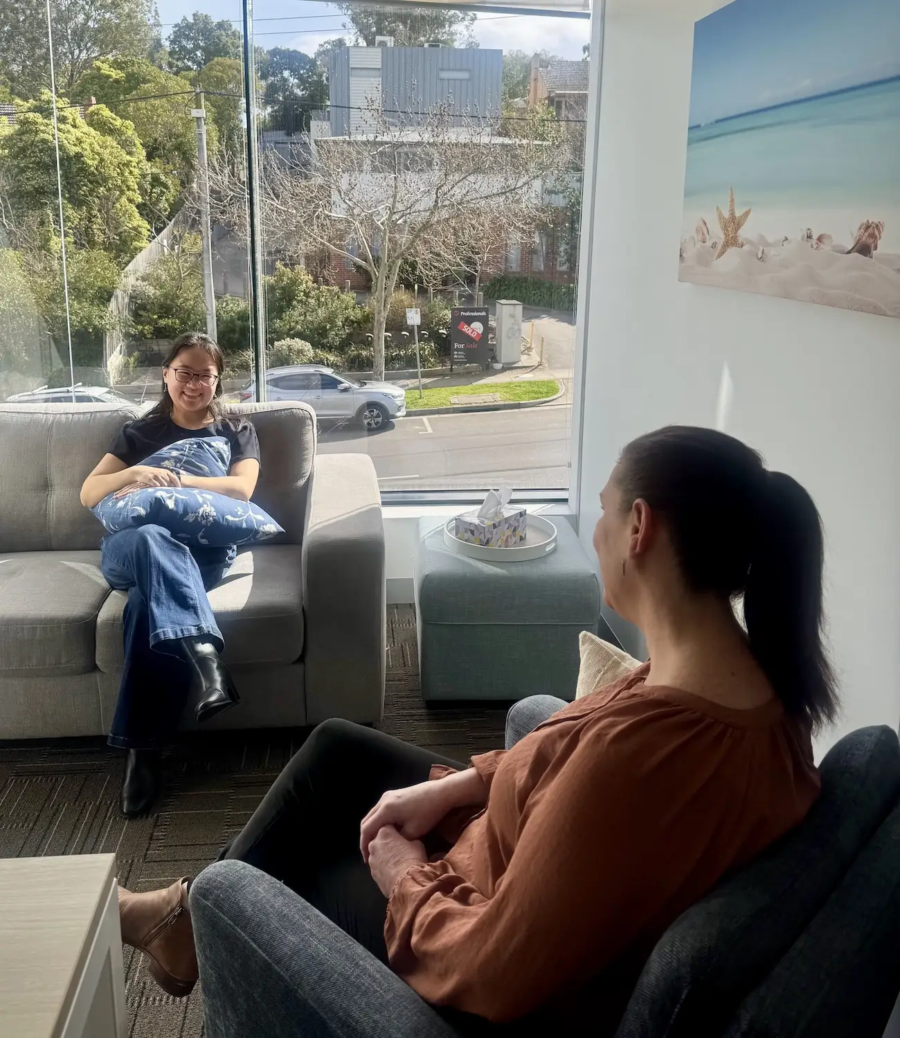 Two women are sitting in a bright room with large windows; one woman is sitting on a gray couch holding a blue floral pillow, smiling, while the other sits in a gray armchair, facing her, wearing a brown top and black pants. Outside the window, trees, cars, and a 'Sold' real estate sign are visible. A box of tissues sits on a teal ottoman next to the window under a beach-themed wall picture.
