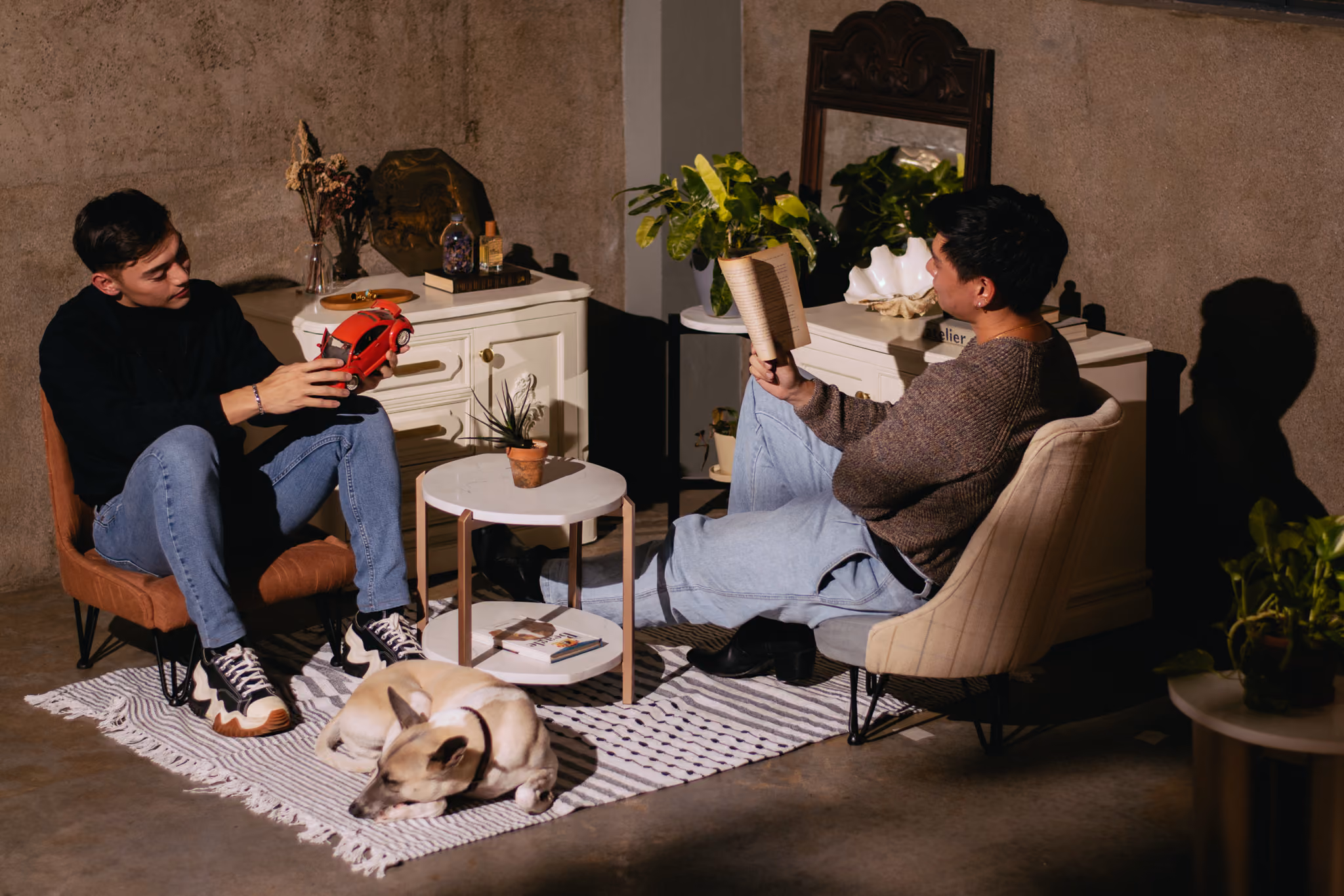Two men sitting indoors with a dog on a rug; one reads a book while the other holds a toy car. T.M. Kawpeng