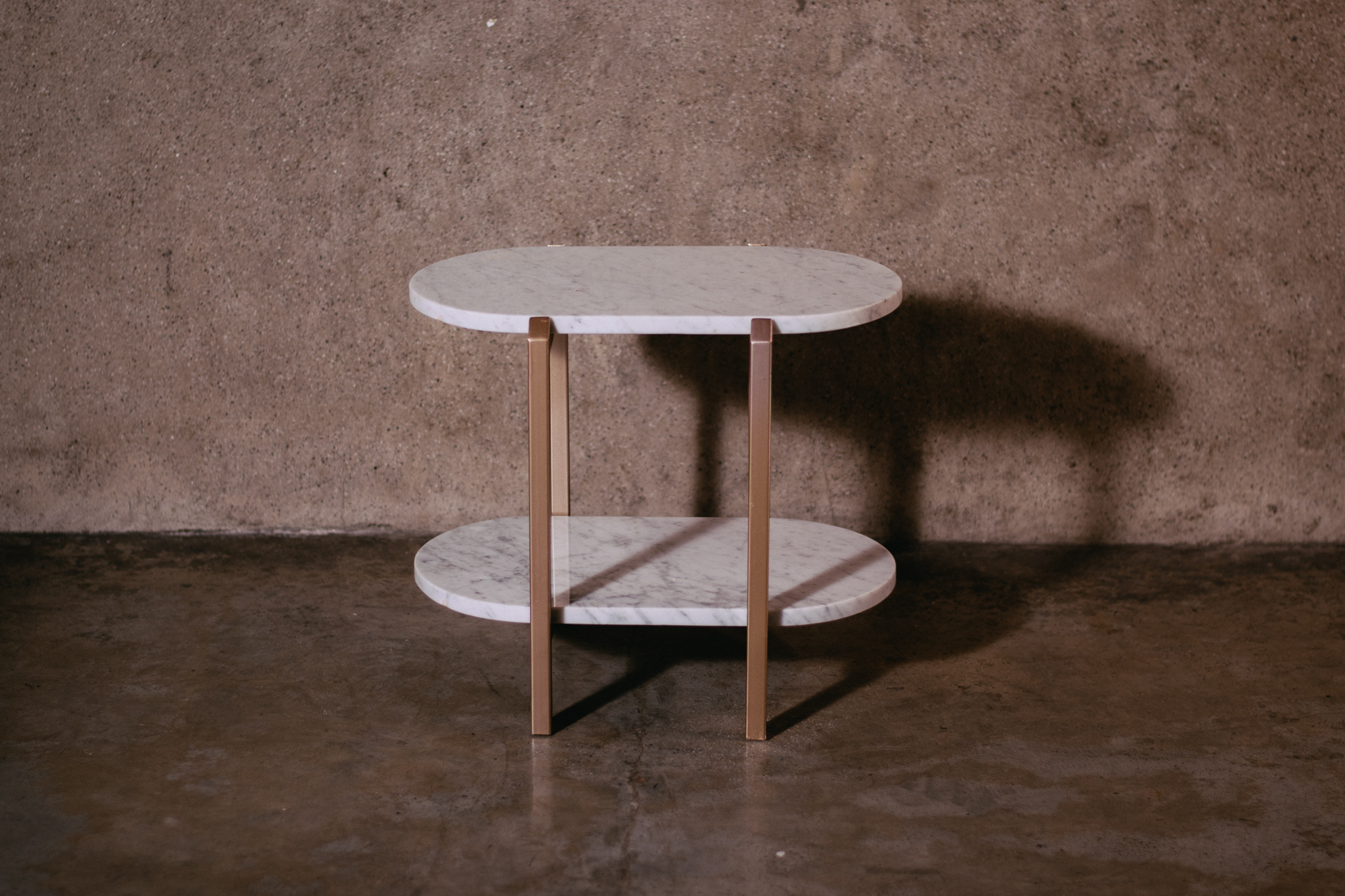 Small two-tier oval white marble table with gold metal legs on a brown floor and wall. T.M. Kawpeng