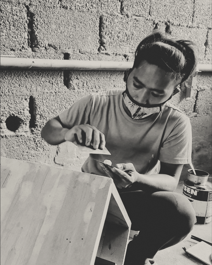 Person wearing face mask sanding or smoothing a wooden object indoors against a brick wall. T.M. Kawpeng