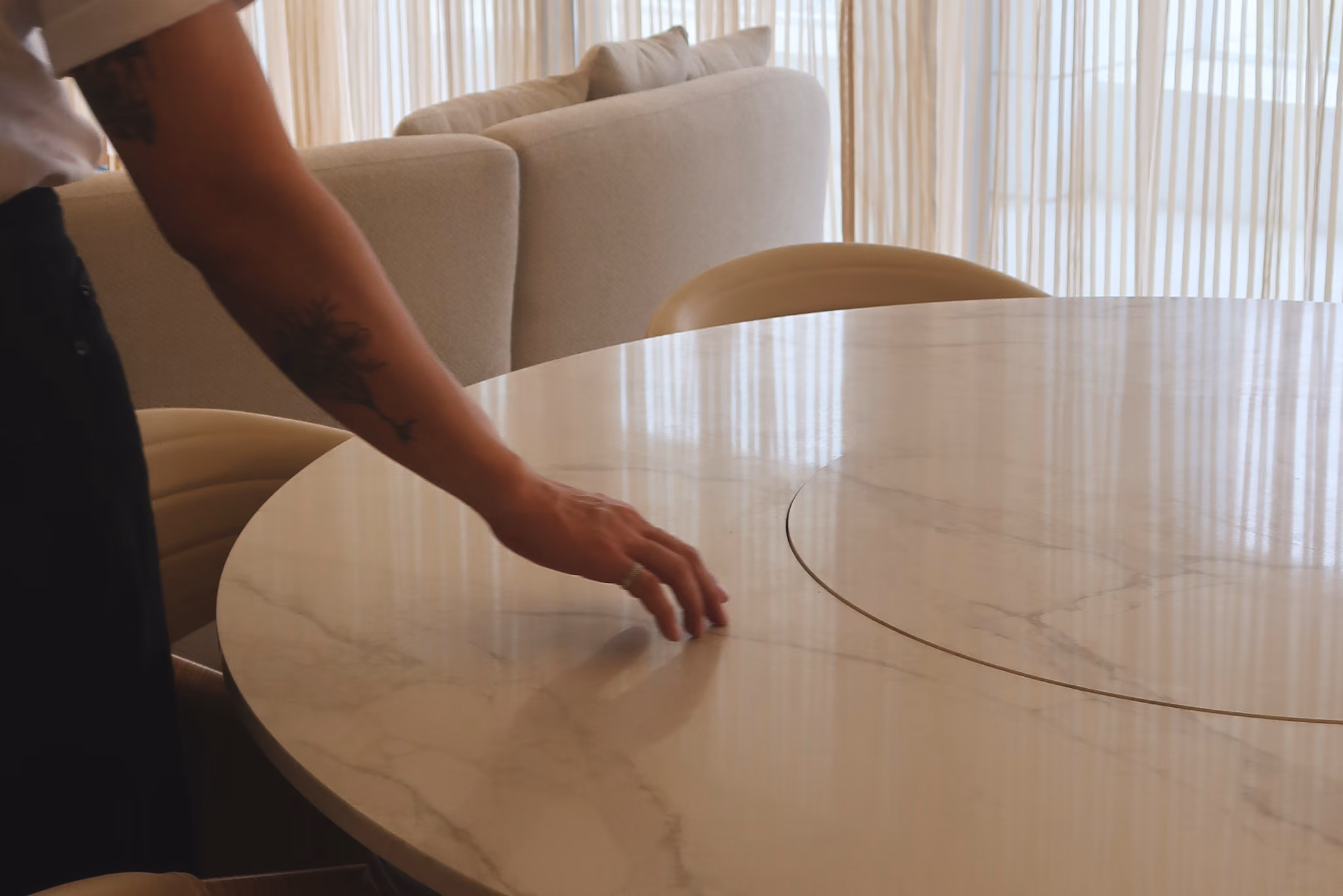 Arm with tattoo reaching across a round marble table in a softly lit room with beige chairs and couch. T.M. Kawpeng