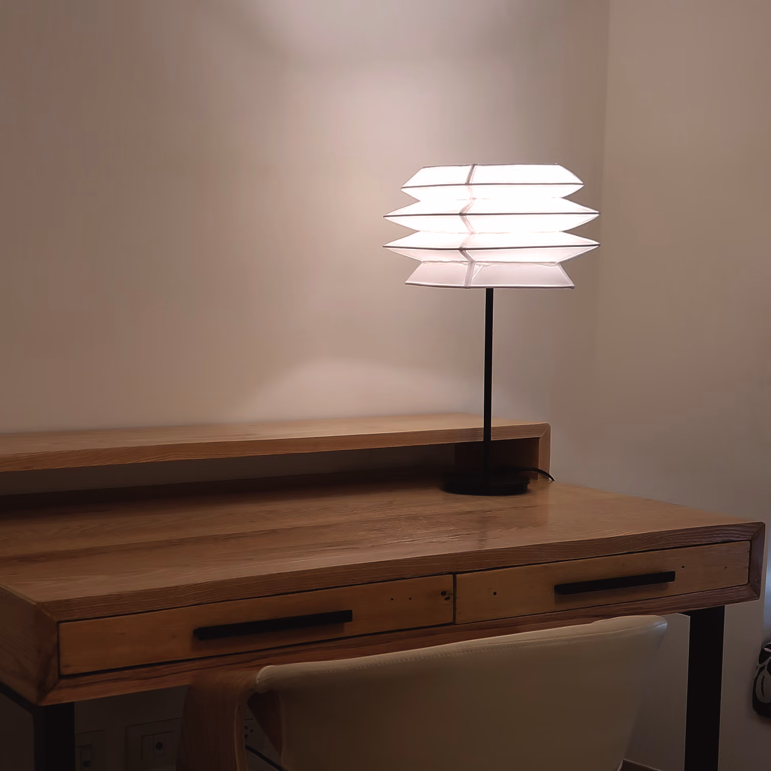Modern desk lamp with layered white shade on a wooden desk with drawers against a plain wall. T.M. Kawpeng