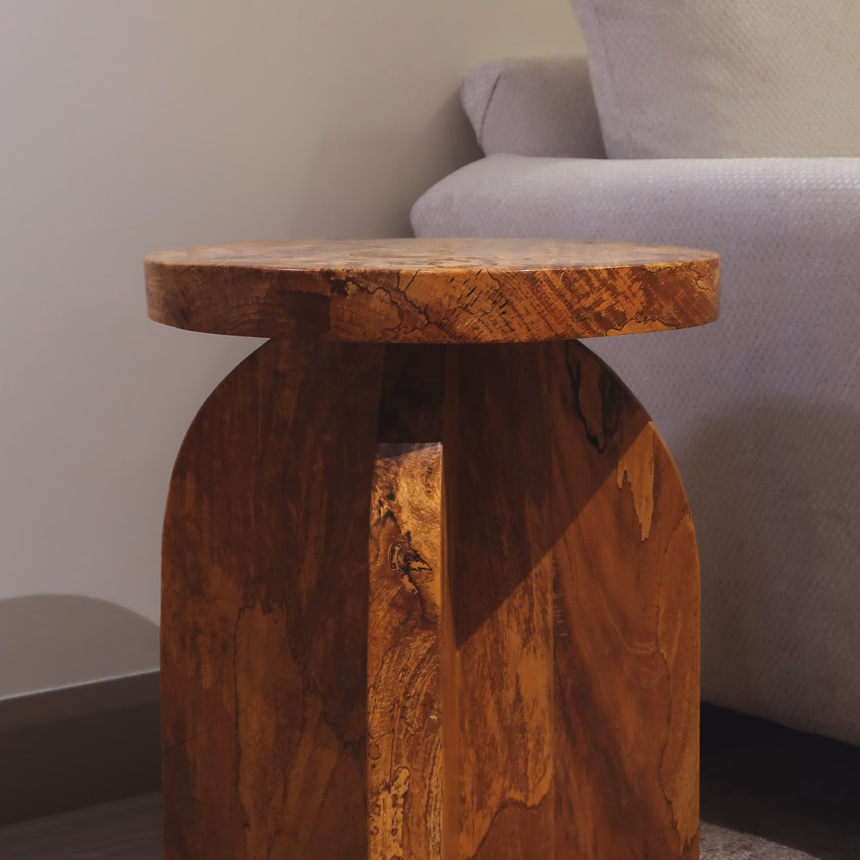 Round wooden side table with natural grain next to a light gray couch. T.M. Kawpeng