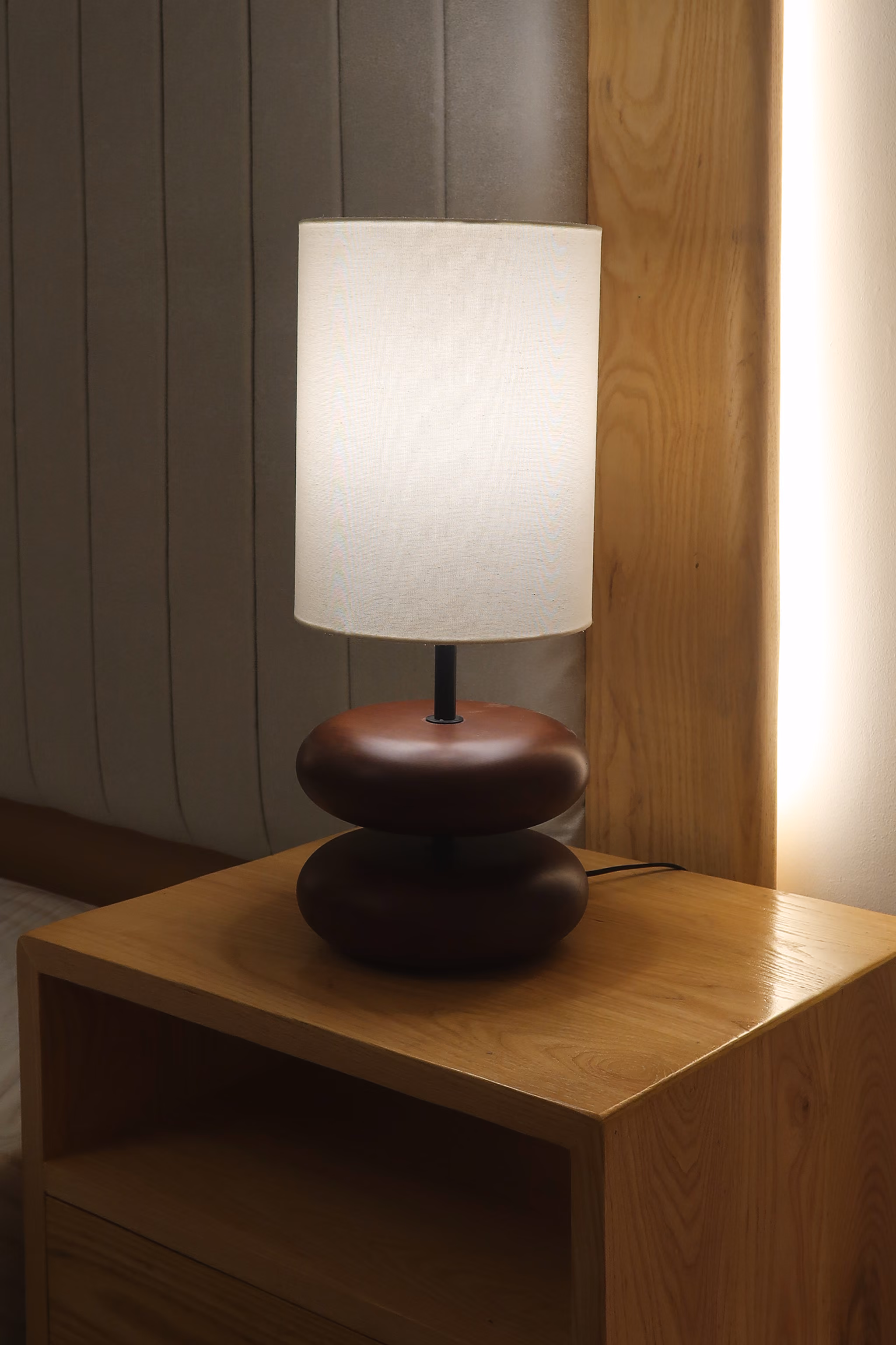 Lit table lamp with white shade and dark brown base on a wooden nightstand next to a bed. T.M. Kawpeng