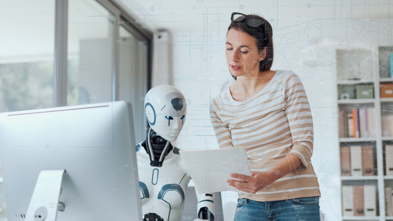 A woman reviewing a document alongside a humanoid robot working at a computer