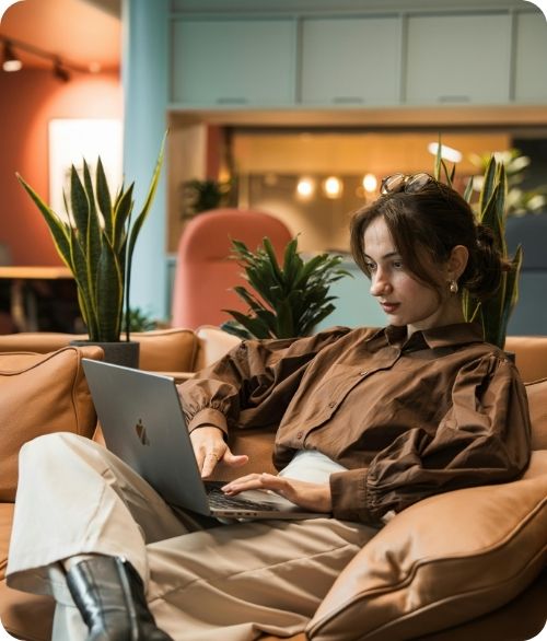 Young woman in brown shirt sitting on couch using laptop with indoor plants around her.