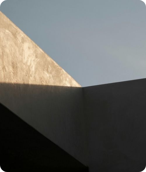 Minimalist architectural corner with sunlight casting shadows on textured walls against a clear sky.