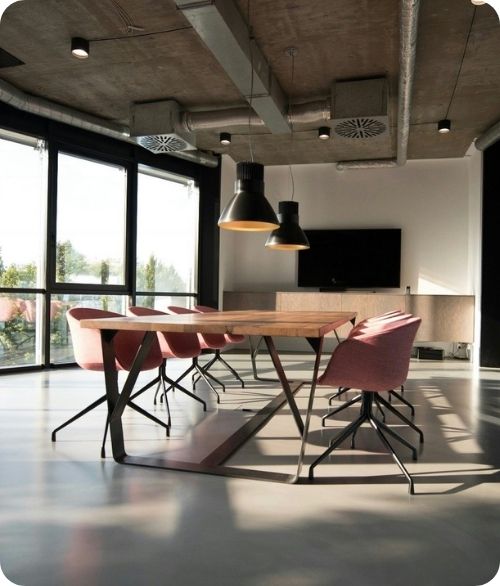 Modern office meeting room with wooden table, pink chairs, large windows, and hanging black lamps.