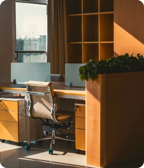 Sunlit modern office workspace with a rolling chair, wooden desks, partitions, and a planter box.