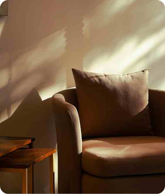 Brown cushioned armchair with a pillow next to nested wooden tables in soft sunlight.