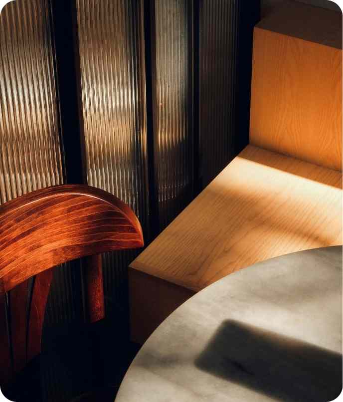 Sunlit wooden chair and bench beside a gray round table by vertical ribbed glass panels.