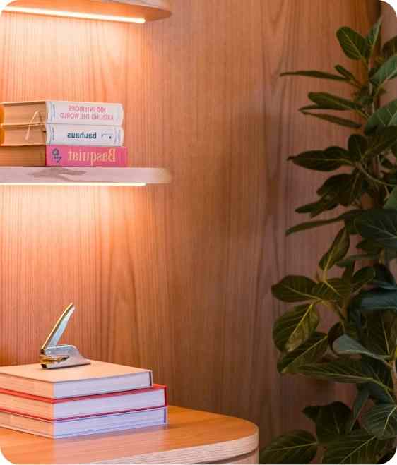 Stacked books and a stapler on a wooden table under a wall-mounted shelf with books and a plant nearby.