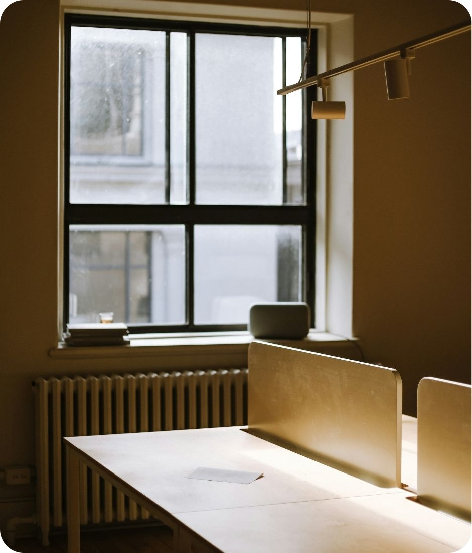 Sunlit modern office workspace with wooden desks separated by beige privacy panels, a large window with black frames in the background, a radiator beneath the window, and a speaker or device on the windowsill.