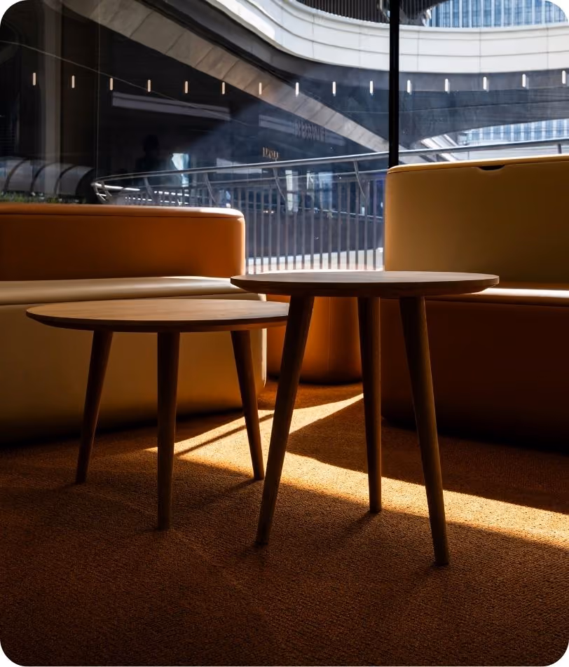 Two round wooden tables with three legs each, placed on a carpeted floor between modern tan leather sofas, with natural light casting shadows from a large window overlooking a cityscape with curved architectural elements.