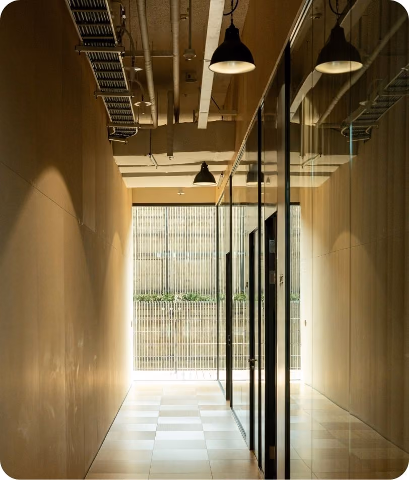 Empty corridor with tiled floor and beige walls, featuring glass doors on the right side and overhead industrial-style lighting.