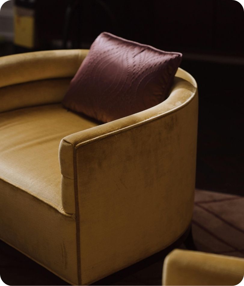 Curved mustard-yellow velvet armchair with a maroon cushion, set against a dark background.