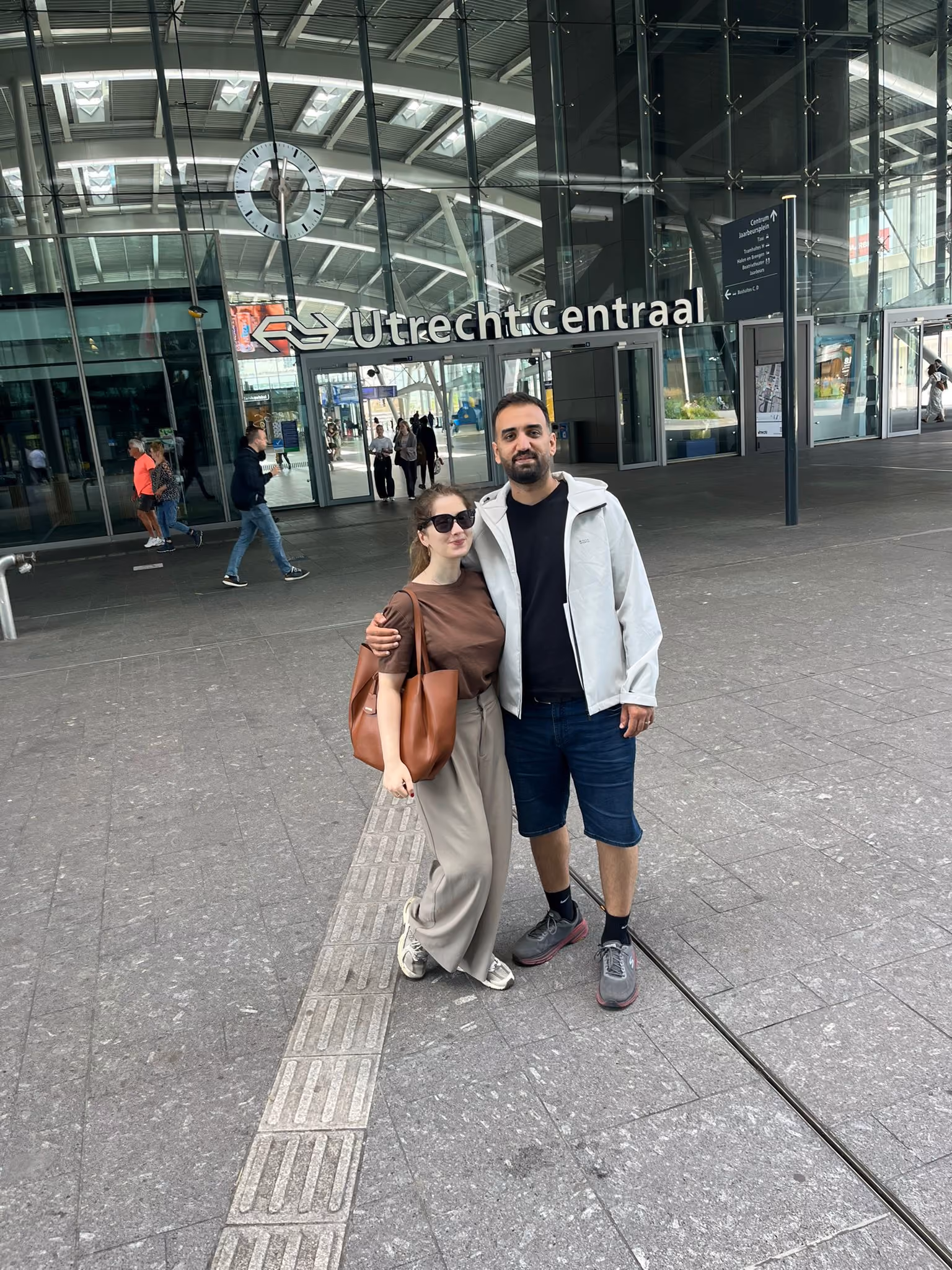 A man and woman standing arm-in-arm outside Utrecht Centraal train station under its glass roof and clock, with a few pedestrians and signage visible in the background.