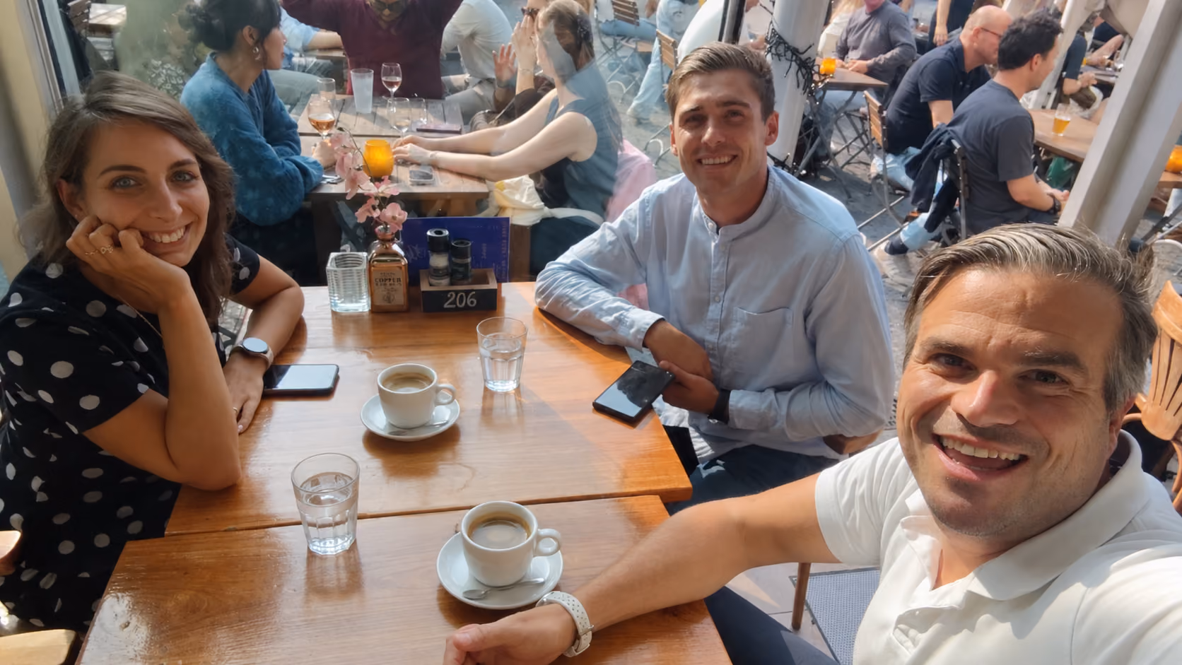 Three people sitting at a wooden table in a cafe with coffee cups and glasses of water, smiling and posing for a selfie taken by the man in a white shirt.