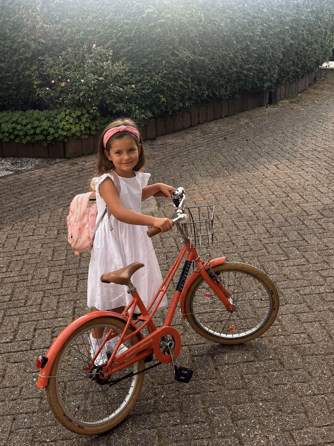 Young girl in white dress with pink headband and backpack standing with a red bicycle on a paved surface near green bushes.
