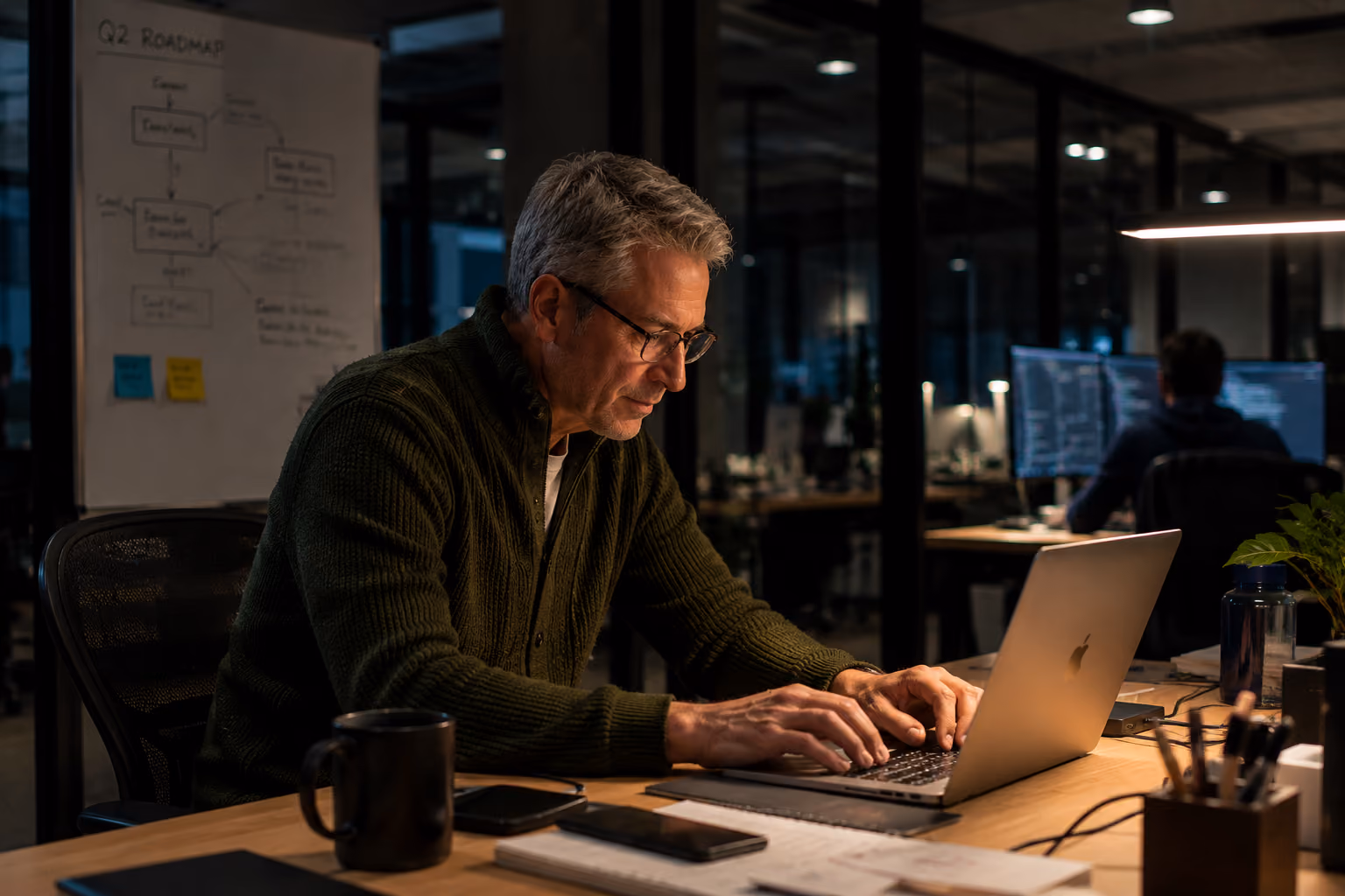 Middle-aged man with glasses working on a laptop at a desk in a dimly lit modern office with another person visible working in the background.