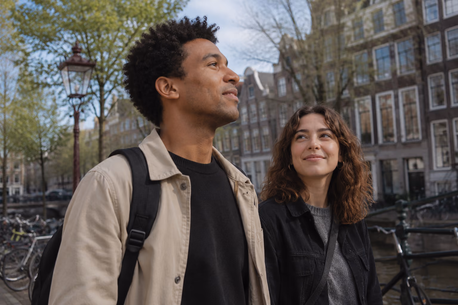 A man and woman walking outdoors by a canal with bicycles and historic buildings in the background, the woman looking at the man with a smile.
