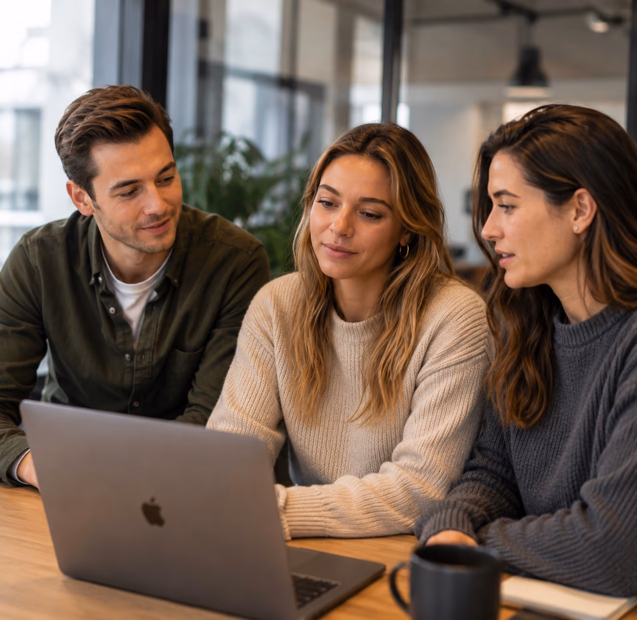 Three young adults sitting at a table looking at a laptop screen, engaged in a discussion inside a modern office space.