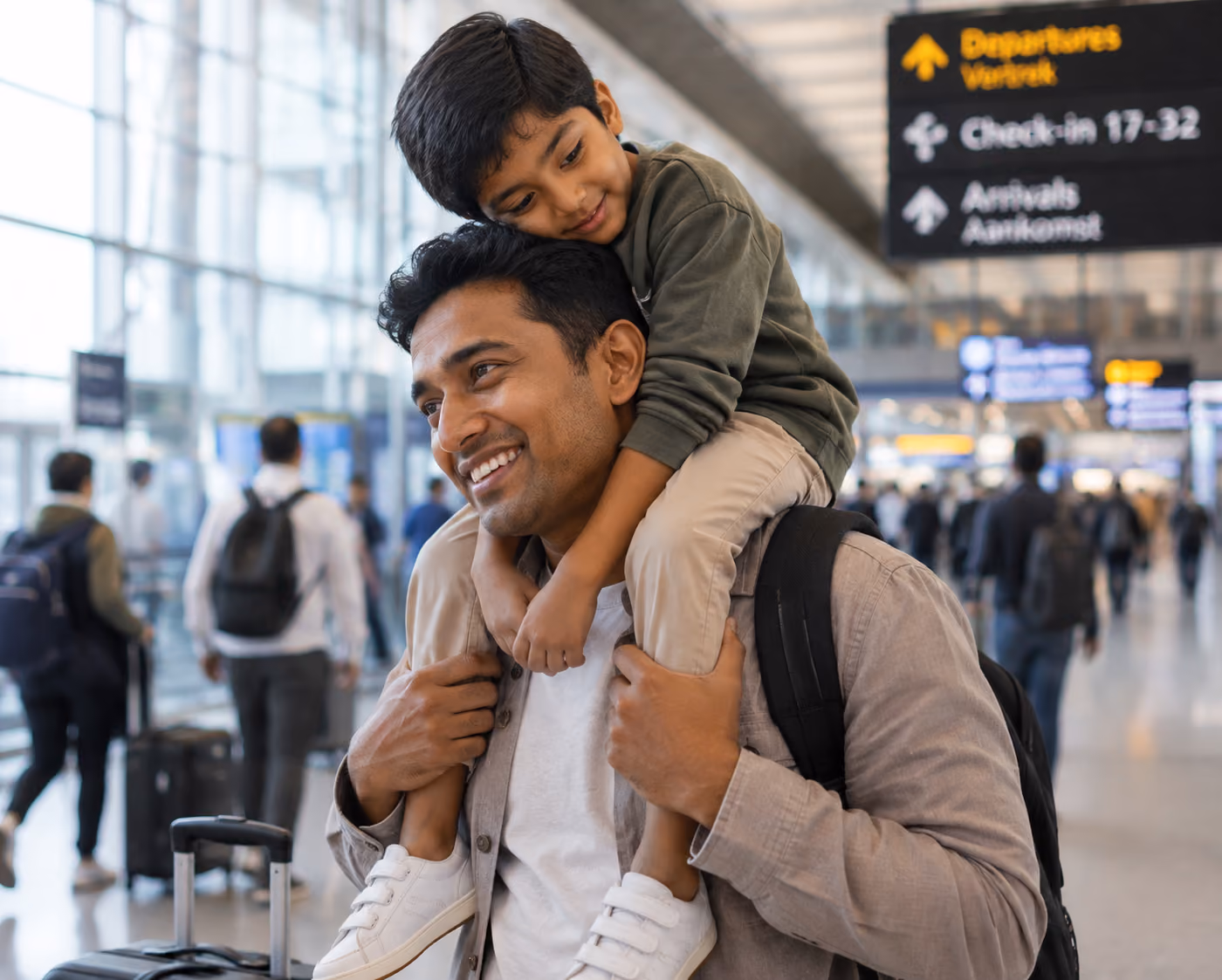Smiling father carrying his son on shoulders in a busy airport terminal, with travelers and directional signs in the background.