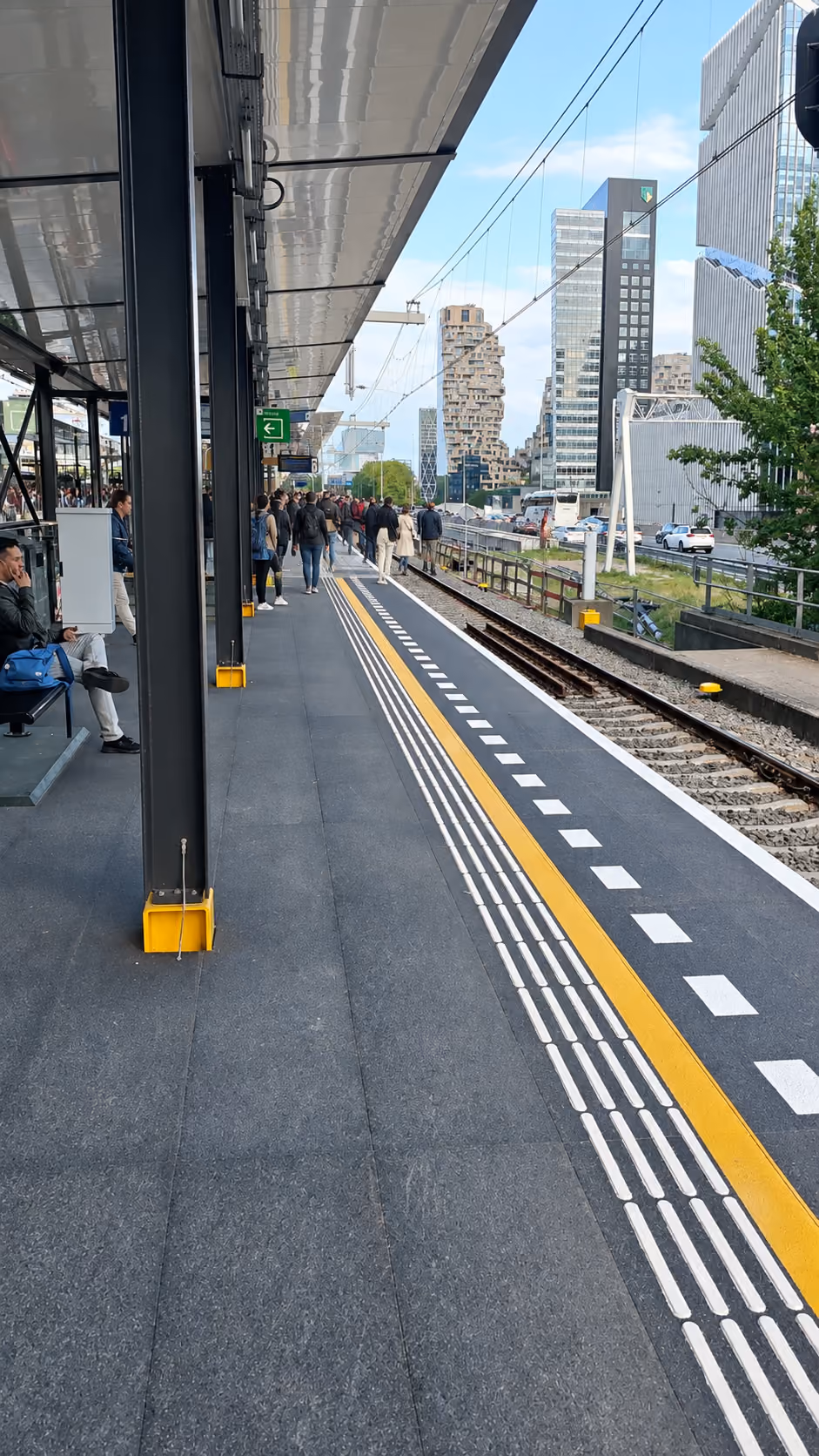 Train station platform with people walking and sitting, tactile paving and yellow safety lines along the tracks, modern city buildings in the background under a partially cloudy sky.