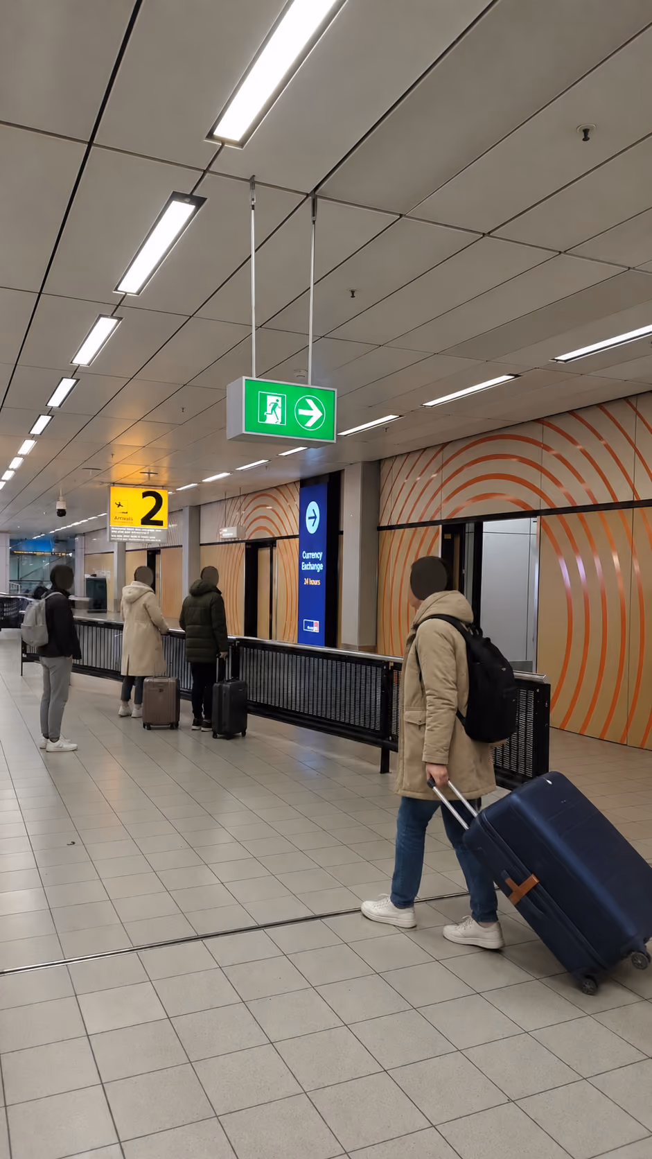 Travelers in coats pull suitcases on wheels through an airport corridor with a green exit sign overhead and a yellow sign indicating gate 2 nearby.